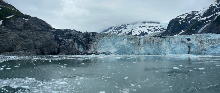 A picture of a large glacier with fissures, hanging over an icy body of water, mountains in the background amongst fog.