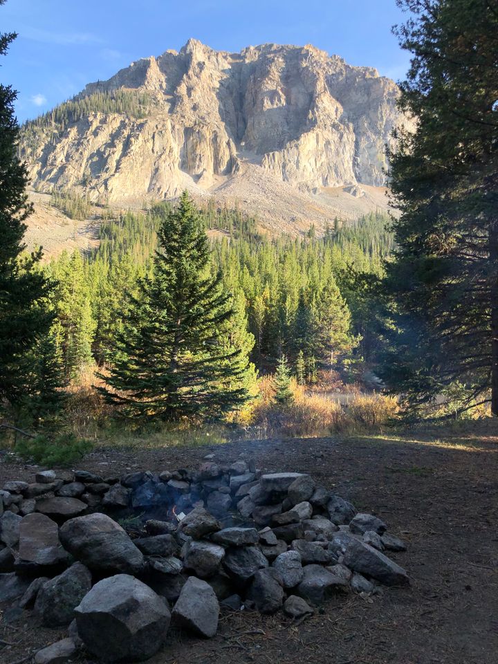 A large jagged mountain in the background, trees in the mid-ground and a campfire smoking in the foreground.